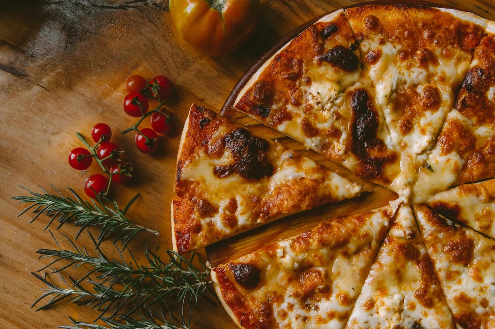 Une pizza au fromage coupée en parts sur une planche en bois, avec des tomates cerises et du romarin